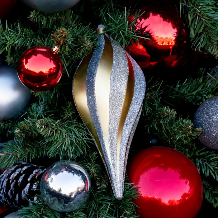 A close-up of a silver and gold striped Christmas ornament nestled among green pine branches and red reflective ornaments.