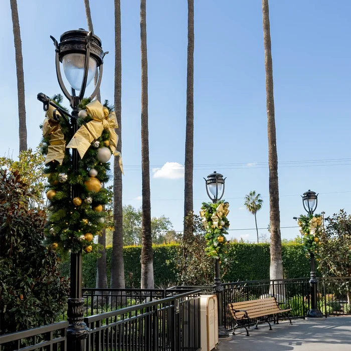 A decorated black lamppost in an outdoor park setting, adorned with a green garland and large gold bows, with tall palm trees, a bench, and other lampposts in the background under a blue sky.