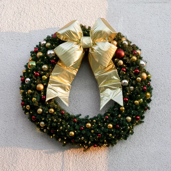 A green Christmas wreath decorated with gold and red ornaments and a large shiny gold bow, hanging against a light textured wall.