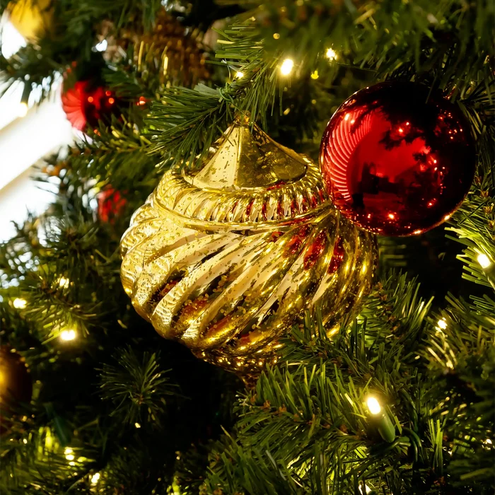 A close-up of a gold ribbed Christmas ornament hanging on a green Christmas tree with warm white lights and a shiny red bauble nearby.