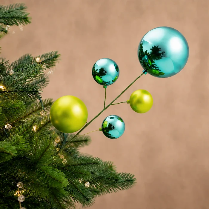 Five shiny Christmas tree ornaments in metallic teal and lime green hang from a thin branch against a warm beige background, with part of a green pine tree visible on the left.