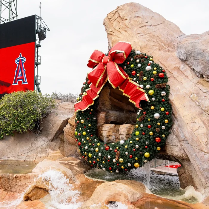 A large Christmas wreath with a red bow and gold trim hangs on a rock formation above a small waterfall. The wreath is decorated with green pine needles and colorful ornaments, and a red Los Angeles Angels stadium sign is visible in the background.