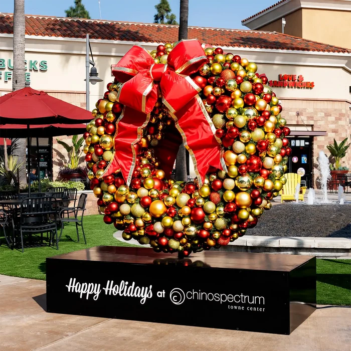 A large festive holiday wreath sculpture covered in red, gold, and silver ornaments, with a oversized red bow, displayed on a black sign base that reads 'Happy Holidays at Chino Spectrum Towne Center' in an outdoor plaza with a restaurant and fountain in the background.