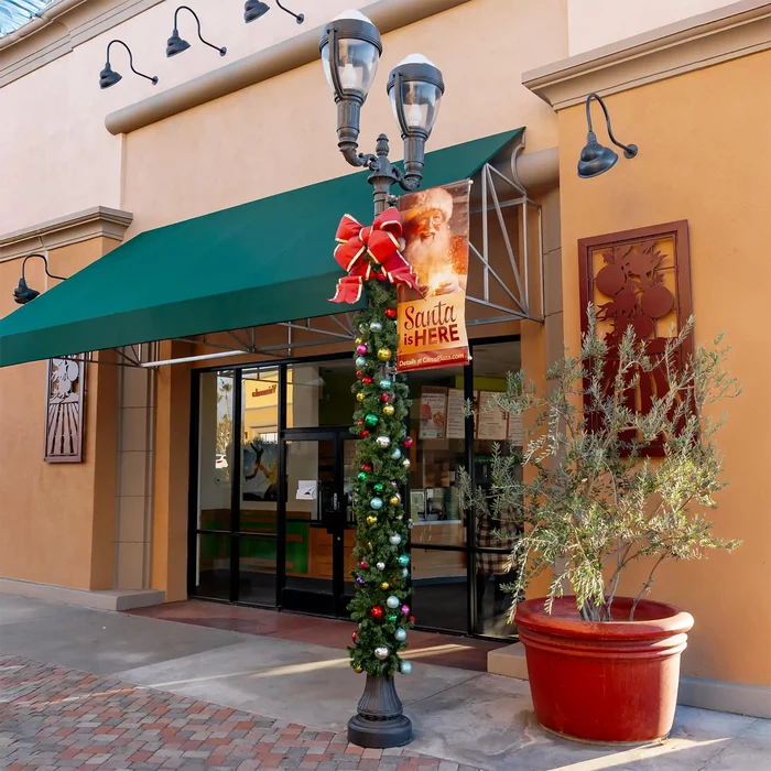 A decorated black double-head lamppost with a Christmas garland, ornaments, and a red structural bow with gold trim stands outside a storefront. The building has a warm tan facade, a green awning, and a large red planter with a leafy plant to the right.
