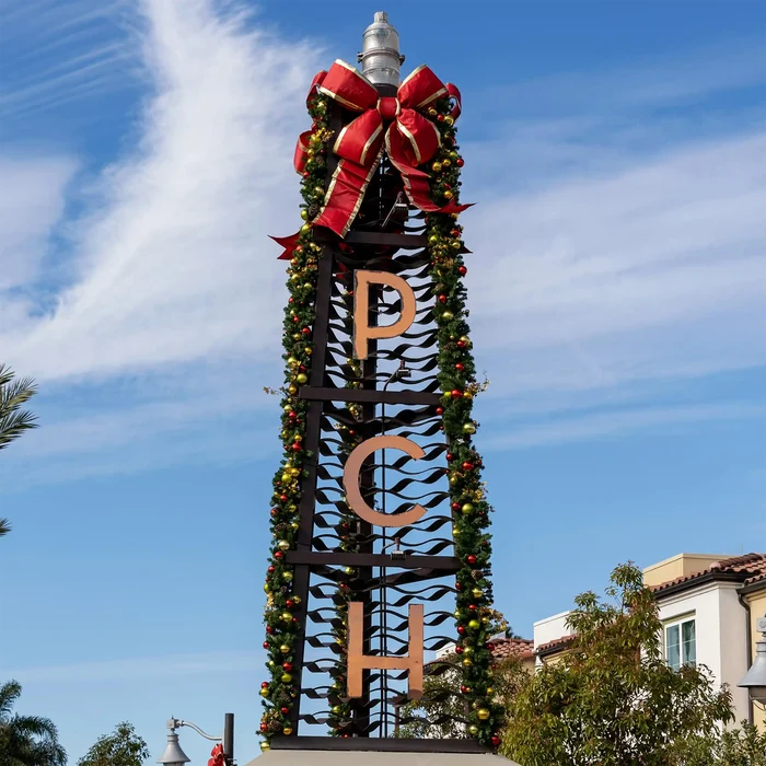 A tall decorative black metal tower outdoors, wrapped in green garland with small ornaments and topped with a large red structural bow with gold trim. The letters PCH are mounted vertically on the tower against a blue sky with clouds.