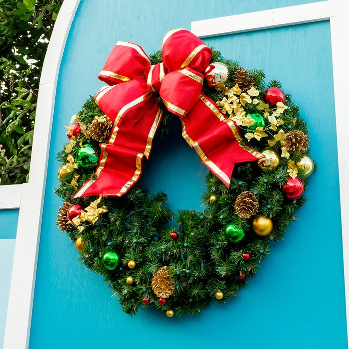 A festive Christmas wreath with a large red bow and gold trim, decorated with green and red ornaments, pinecones, and gold stars, hanging on a bright blue door.